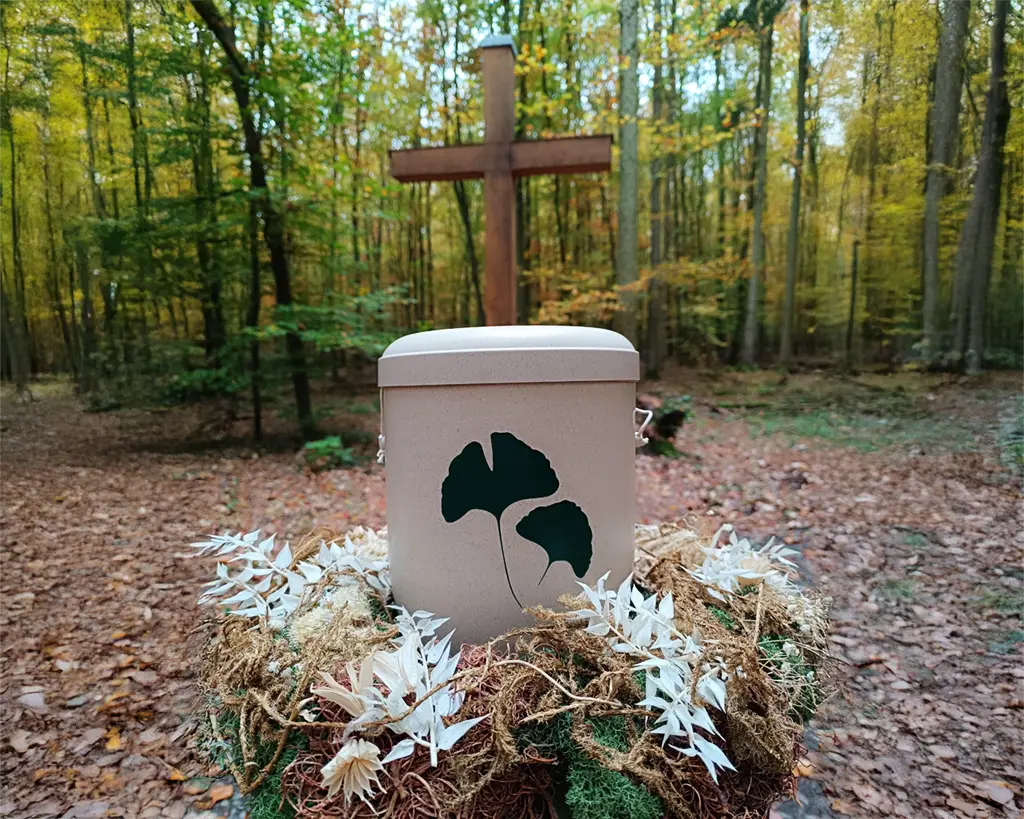 Eine Urne ist mit Blumen verziert und steht auf dem Waldboden vor einem Kreuz aus Holz
